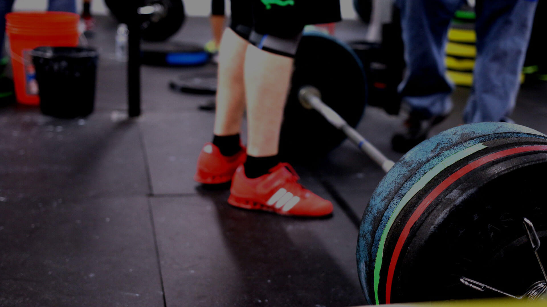 A pair of legs and feet wearing tennis shoes standing next two a barbell with heavy weight